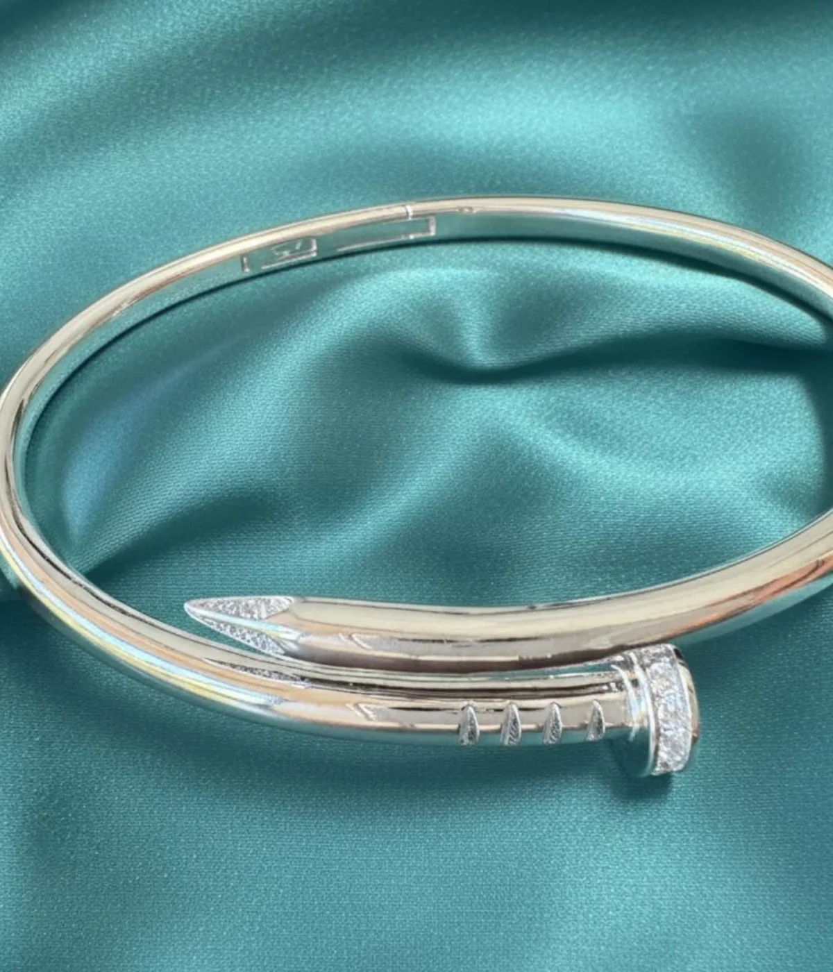 Men's Sterling Silver Nail Kada (cuff bracelet). The sleek, modern design features pave-set Cubic Zirconia stones detailing the nail head and tip. Photographed on a luxurious teal satin surface.