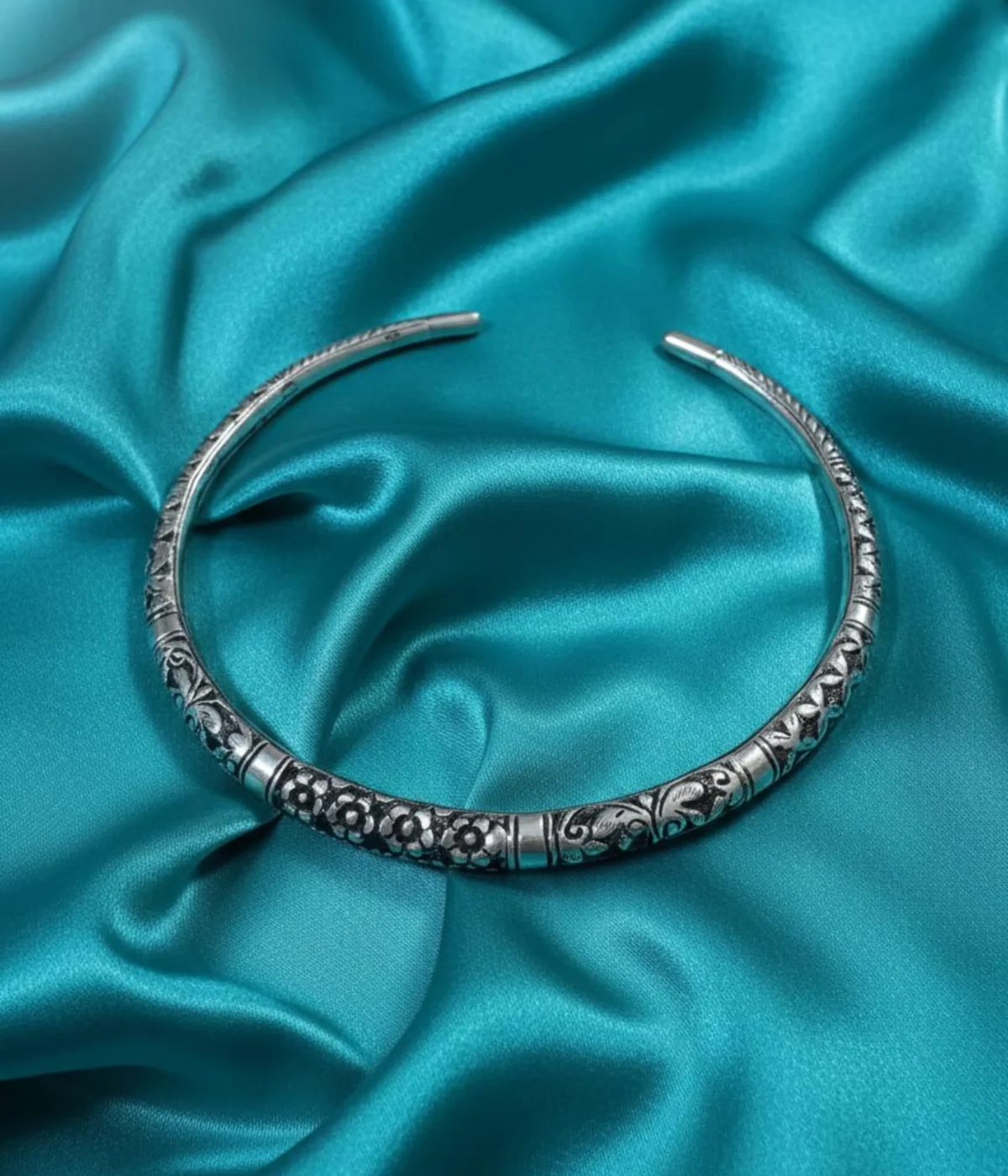 Oxidized Silver Floral Choker Necklace. Rigid, open cuff collar with detailed tribal engraving and an antique finish, displayed on ruffled teal silk fabric.