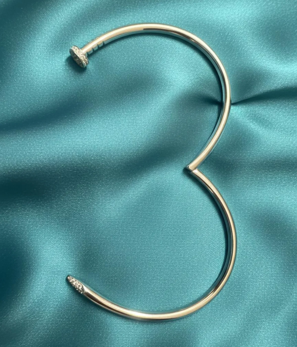 Men's Sterling Silver Nail Kada (cuff bracelet). The sleek, modern design features pave-set Cubic Zirconia stones detailing the nail head and tip. Photographed on a luxurious teal satin surface.