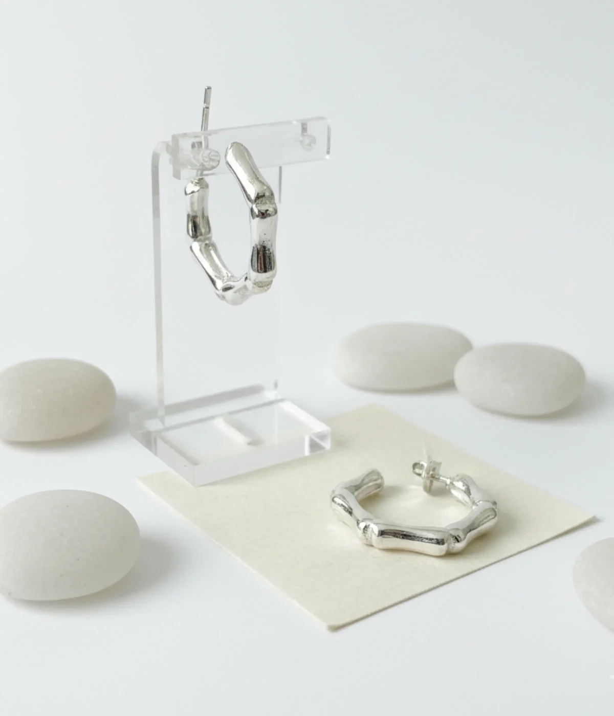 Pair of Silver Bamboo Hoop Earrings. Small, chunky hexagon shape with a segmented, organic textured finish, displayed on white paper next to driftwood and sea glass.