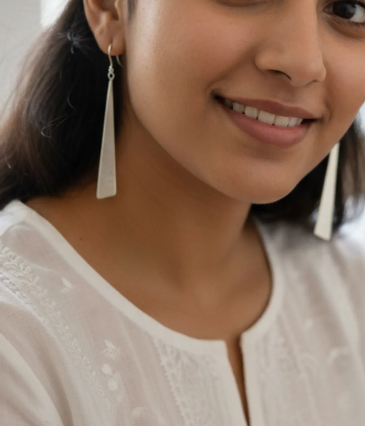 Minimalist Silver Triangle Drop Earrings. Sleek, long geometric dangles on ear hooks, displayed on textured white paper and teal silk, surrounded by smooth green sea glass.