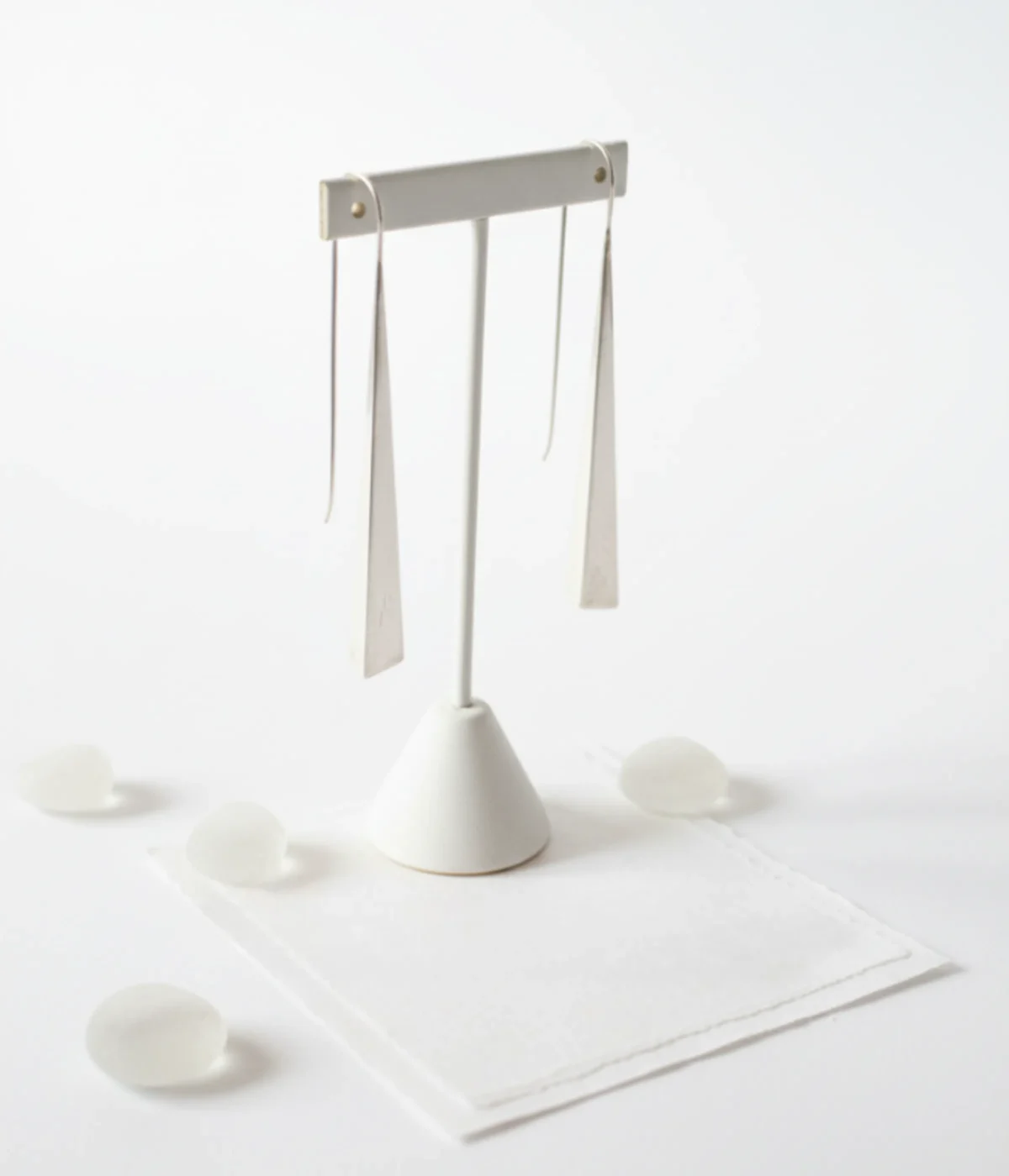 Minimalist Silver Triangle Drop Earrings. Sleek, long geometric dangles on ear hooks, displayed on textured white paper and teal silk, surrounded by smooth green sea glass.