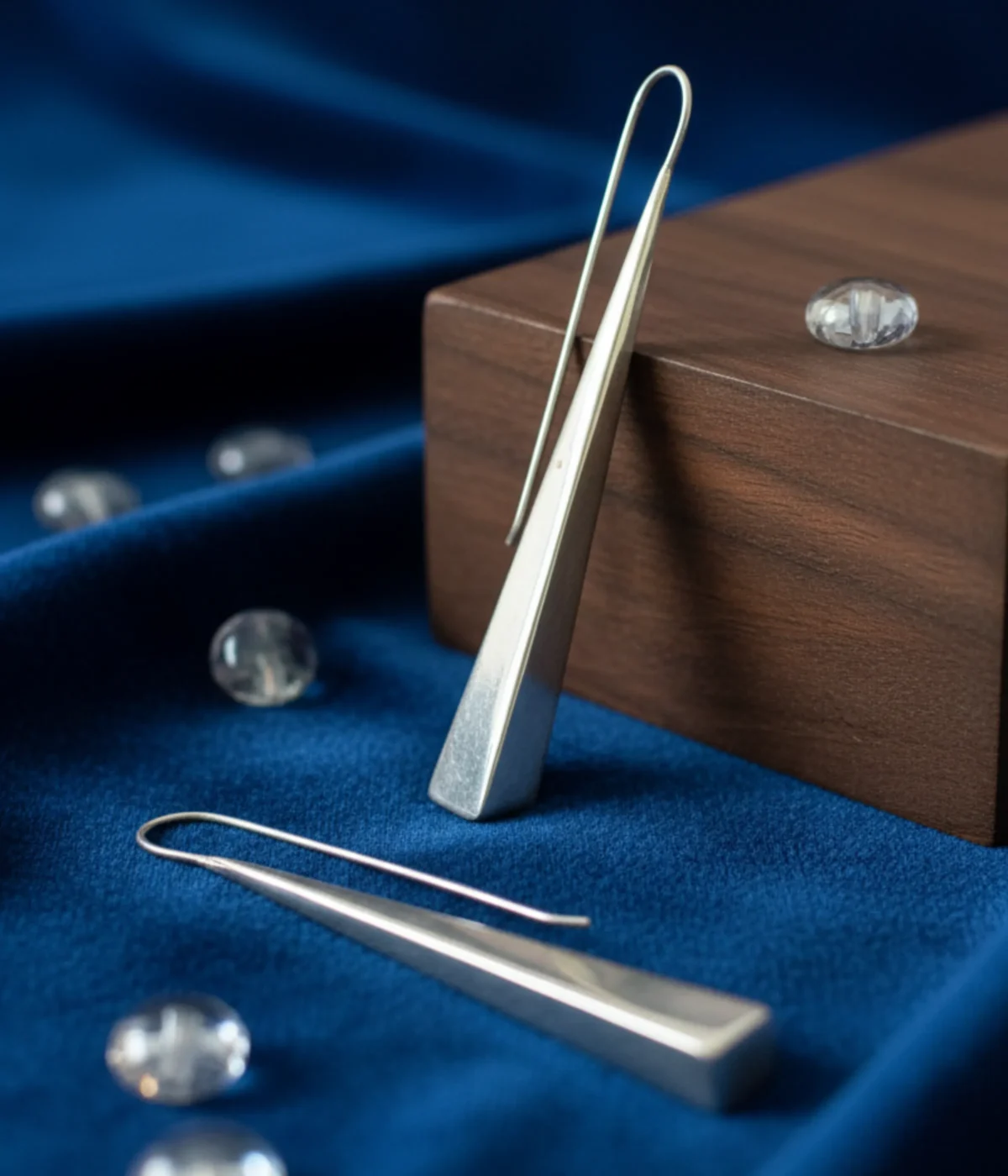 Minimalist Silver Triangle Drop Earrings. Sleek, long geometric dangles on ear hooks, displayed on textured white paper and teal silk, surrounded by smooth green sea glass.