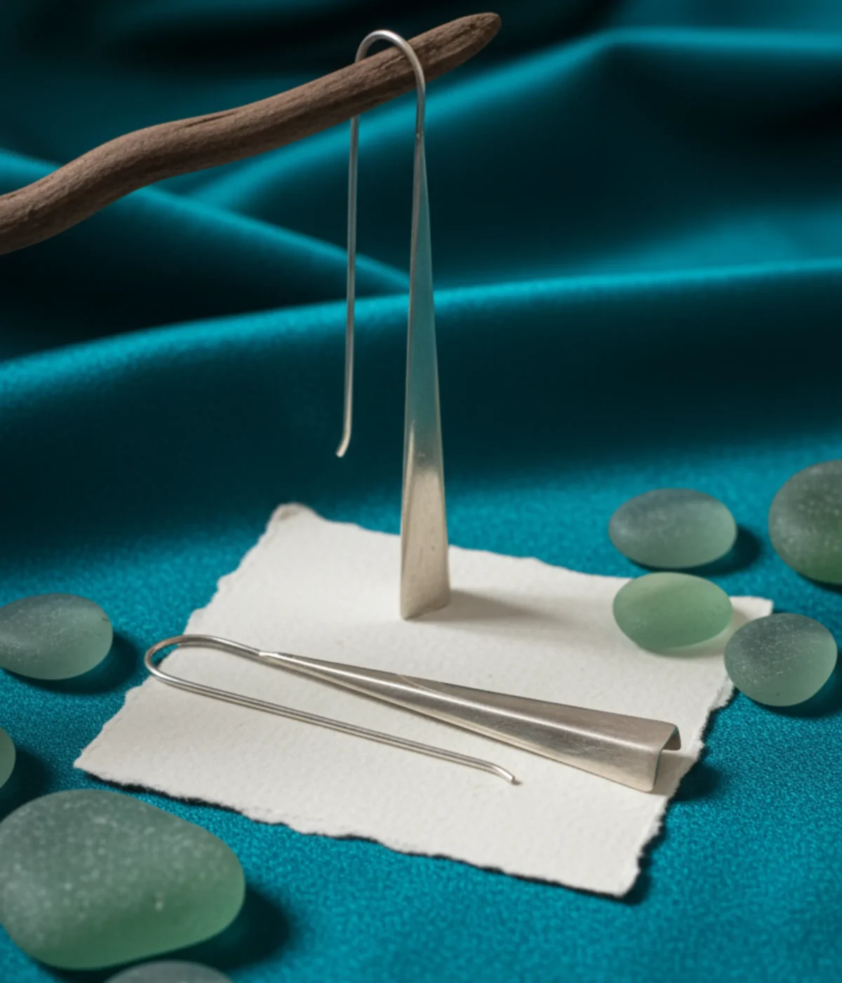 Minimalist Silver Triangle Drop Earrings. Sleek, long geometric dangles on ear hooks, displayed on textured white paper and teal silk, surrounded by smooth green sea glass.