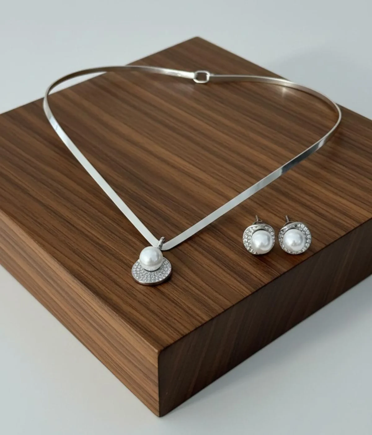 Sterling Silver Collar Necklace and Pearl Stud Earring Set. Features a sleek silver band, a Zircon pave pendant, and matching pearl stud earrings displayed on a wooden riser.