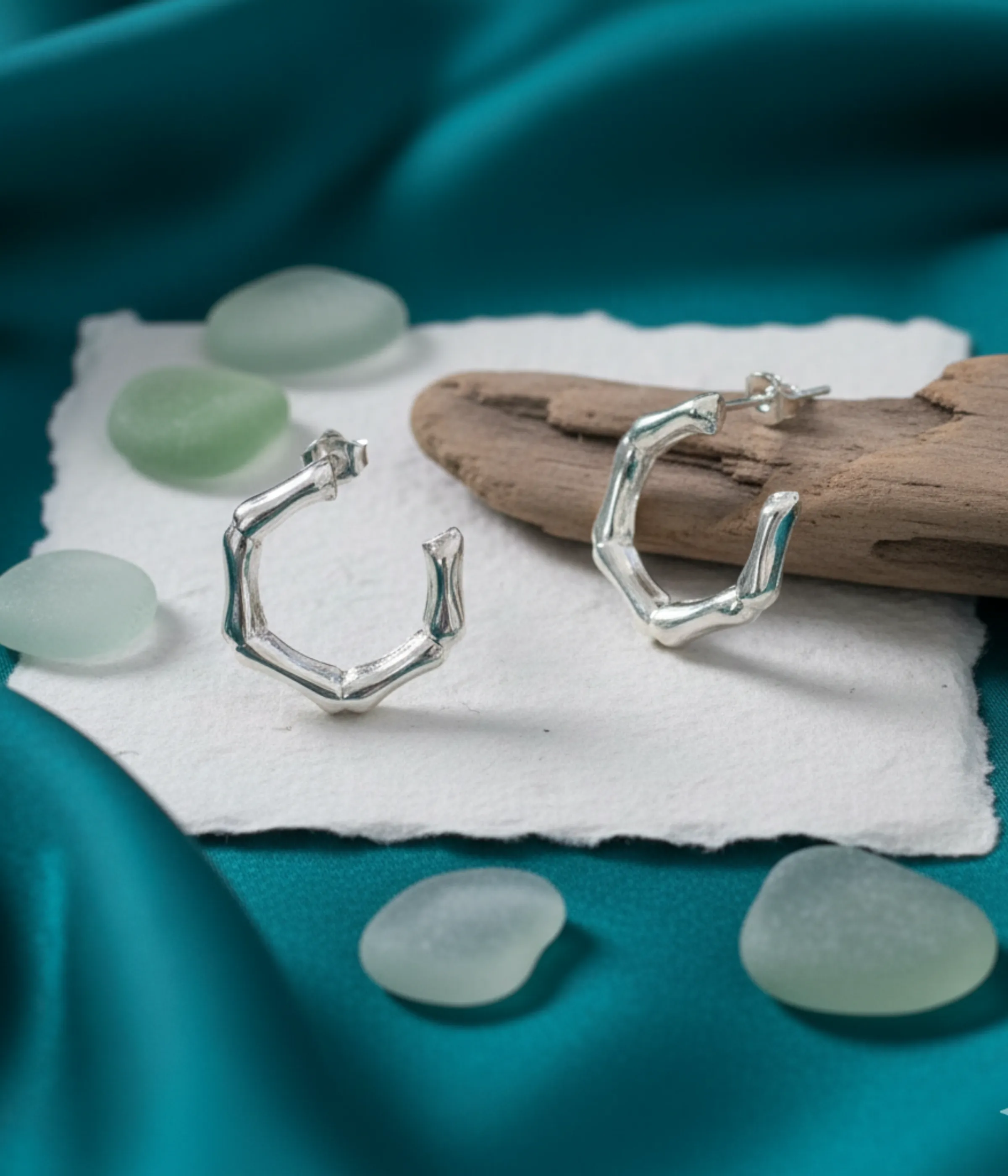 Pair of Silver Bamboo Hoop Earrings. Small, chunky hexagon shape with a segmented, organic textured finish, displayed on white paper next to driftwood and sea glass.