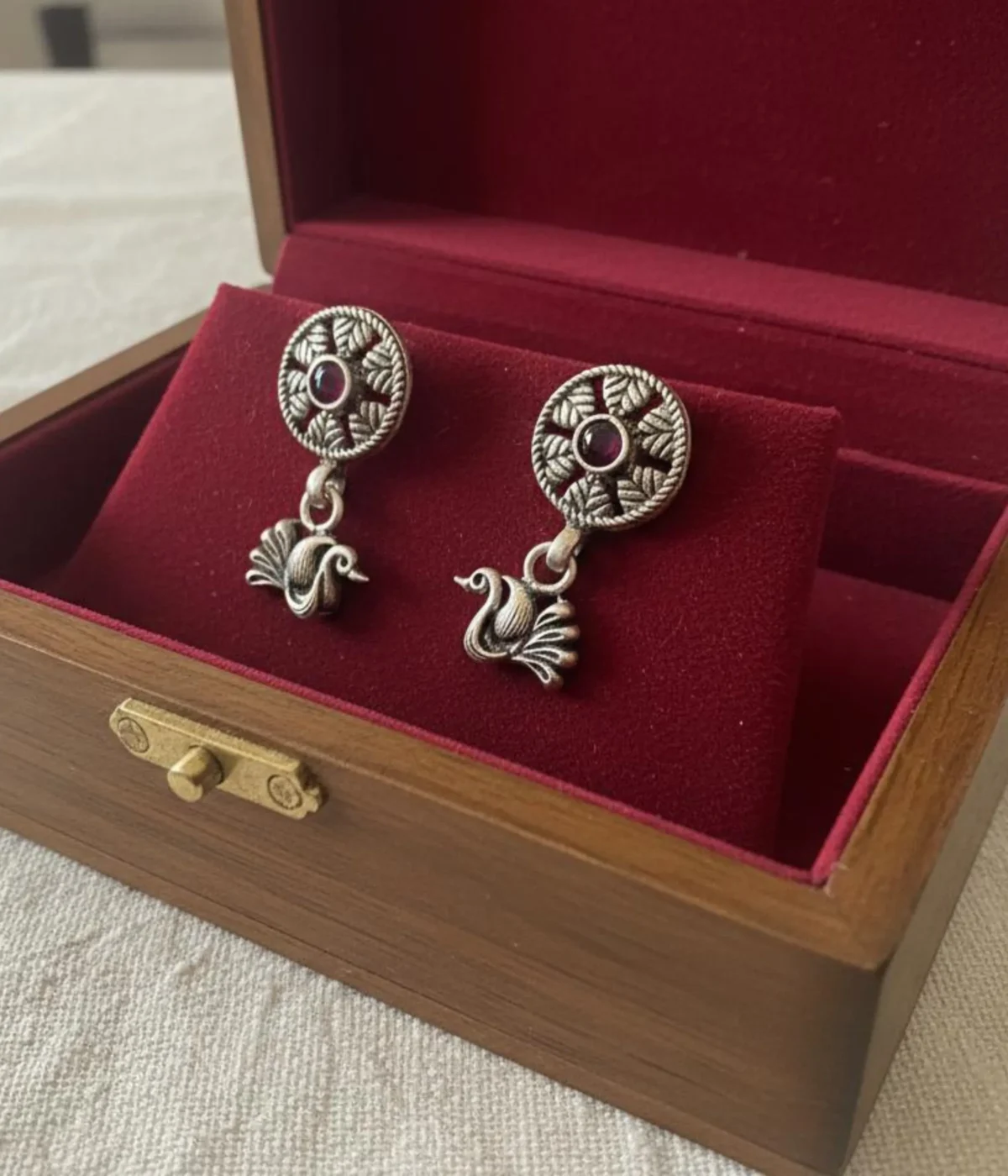 Pair of Oxidized Silver Peacock Drop Earrings. Features a central ruby stone stud and a detailed peacock dangle, displayed on red velvet inside a wooden jewelry box.