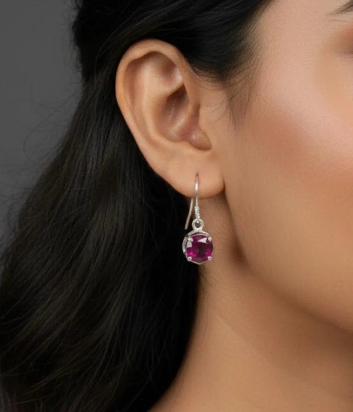 Pair of Minimalist Round Ruby Stone Hook Earrings (1 gm). Features a pink/ruby solitaire stone in a silver six-prong setting, displayed on a red velvet stand inside a dark wood gift box.