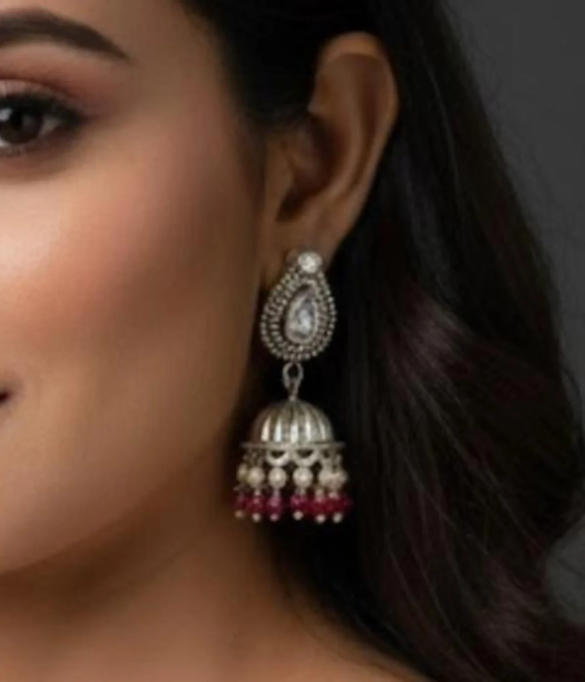 Traditional Oxidized Silver Jhumka Earrings (13 gm) being worn. Features a clear teardrop stone top, bell shape, and contrasting Red Bead Drop Dangles. Classic Indian festive jewelry.