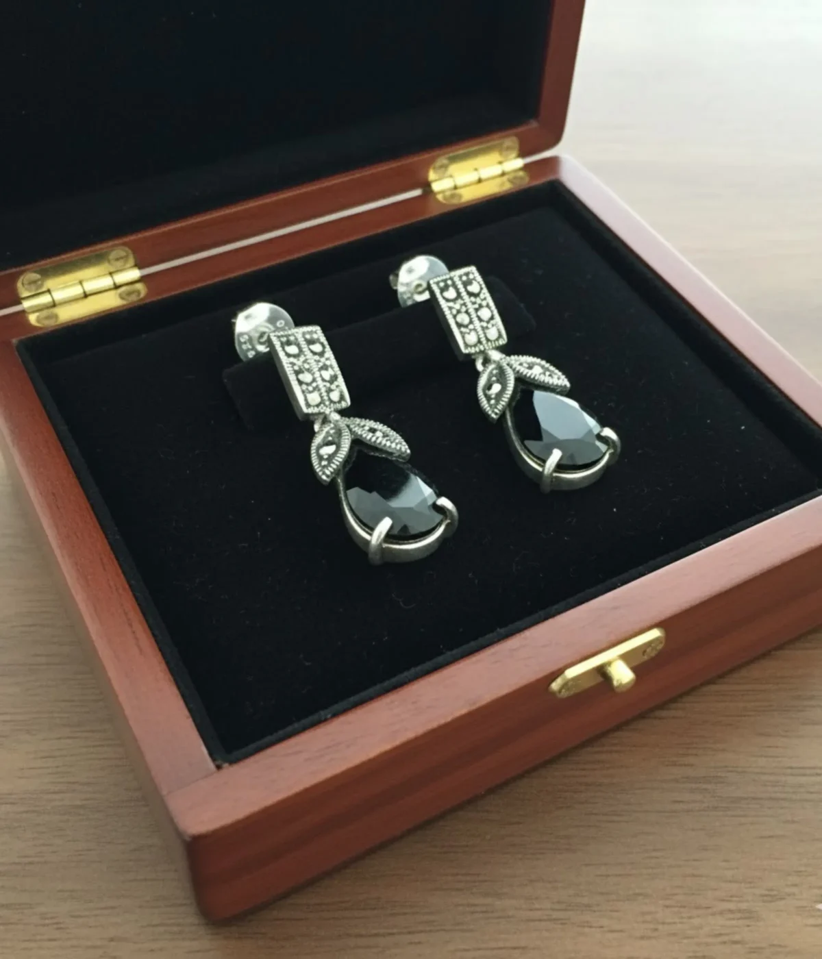 Oxidized Silver Black Onyx Drop Earrings (6.3 gm) with ornate vintage detailing and a teardrop black stone. The earrings are shown resting on a blue velvet pillow inside a wooden jewelry box on a vanity table.