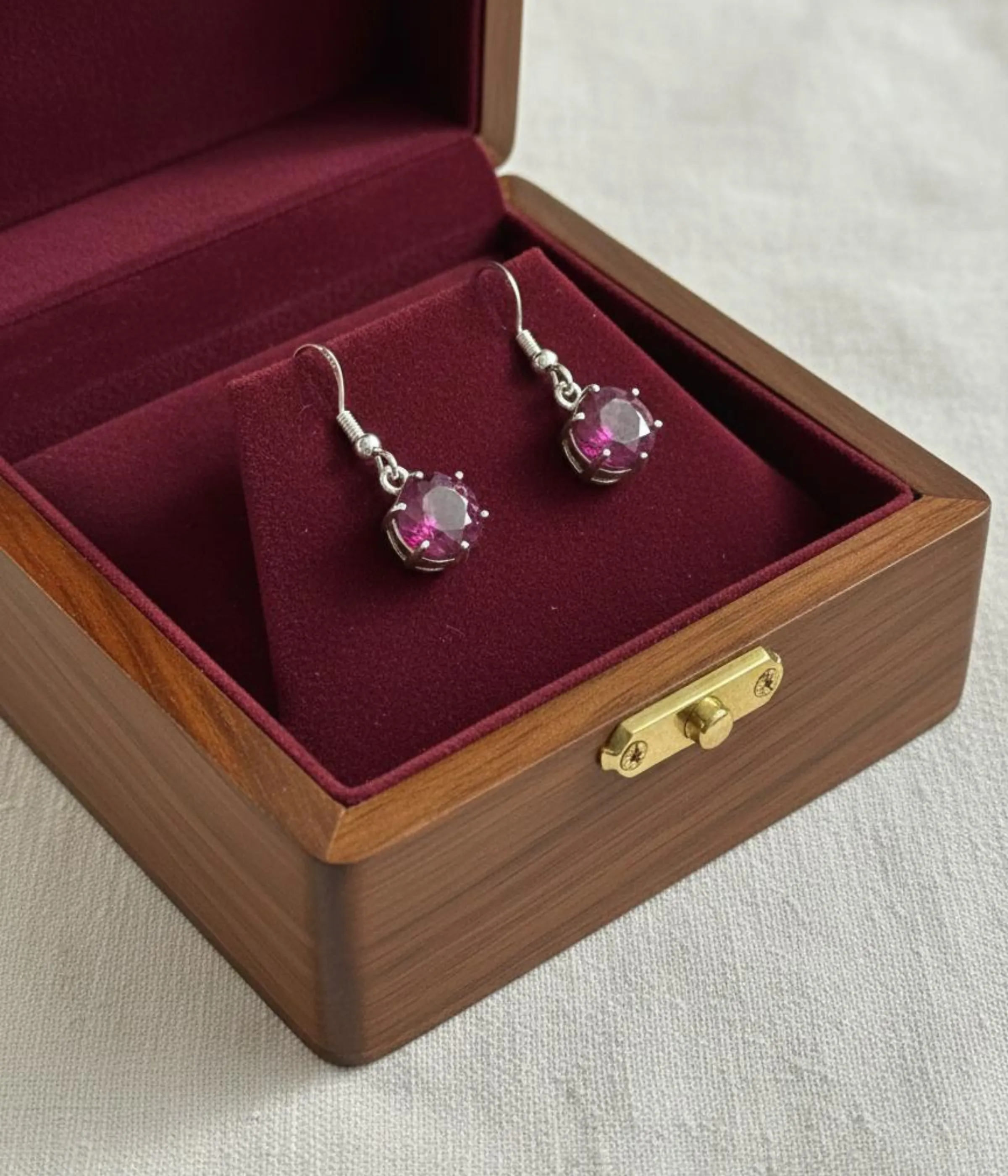 Pair of Minimalist Round Ruby Stone Hook Earrings (1 gm). Features a pink/ruby solitaire stone in a silver six-prong setting, displayed on a red velvet stand inside a dark wood gift box.