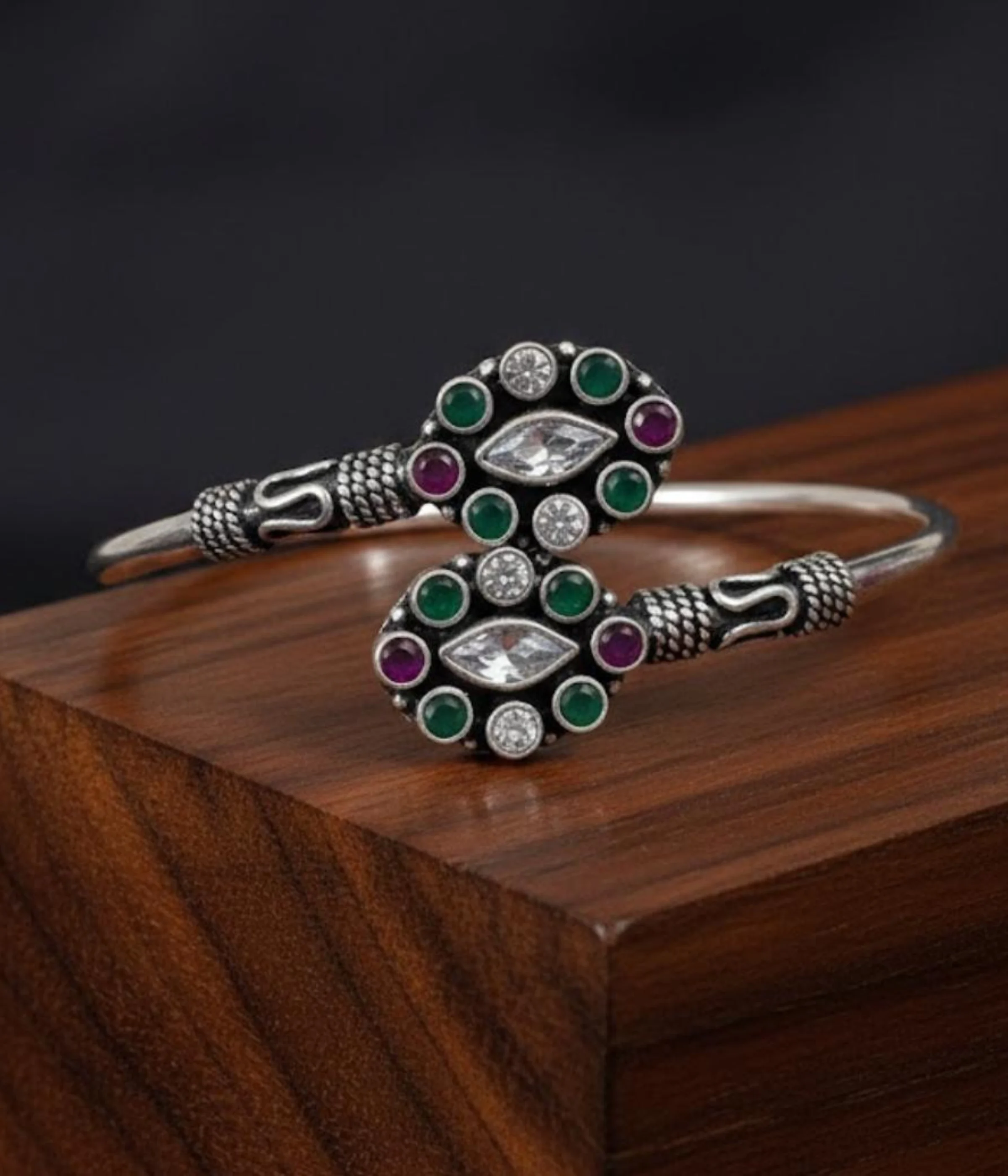 Detailed 16.3 gm oxidized silver floral kada featuring emerald green and maroon stones with clear zirconia accents on a wooden texture.