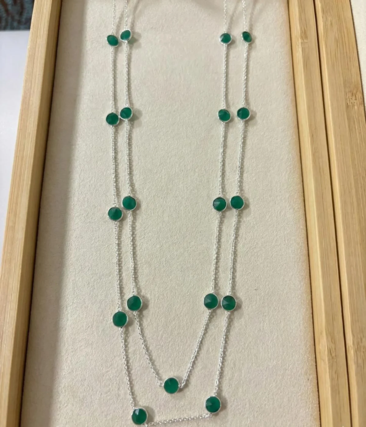 A long 925 sterling silver station necklace featuring vibrant green round gemstones set in polished silver bezel frames, elegantly displayed on a neutral fabric background within a wooden jewelry tray.