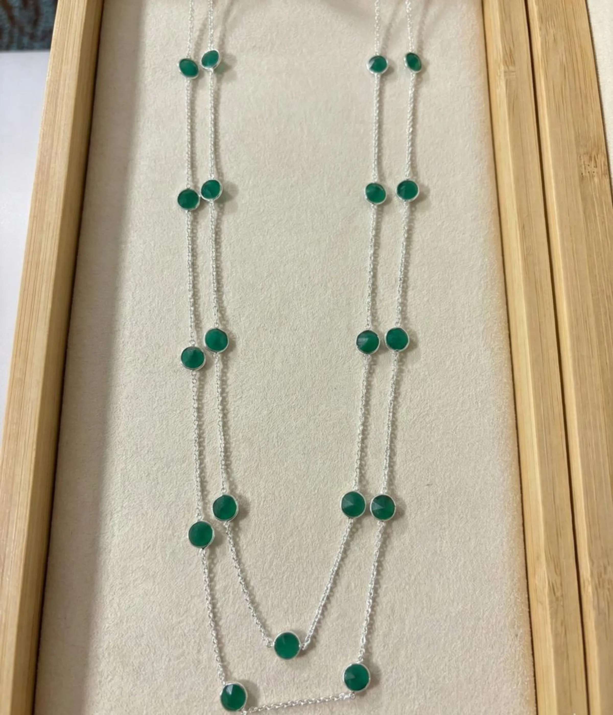 A long 925 sterling silver station necklace featuring vibrant green round gemstones set in polished silver bezel frames, elegantly displayed on a neutral fabric background within a wooden jewelry tray.