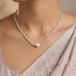 A delicate silver-plated necklace with a shimmering, textured chain and a single, central white pearl, displayed inside a wooden jewelry box on a soft cream-colored cushion.