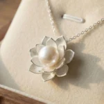 A close-up shot of a silver-plated lotus flower pendant featuring intricate, brushed-metal petals layered around a single, luminous white pearl, displayed on a soft cream jewelry cushion.