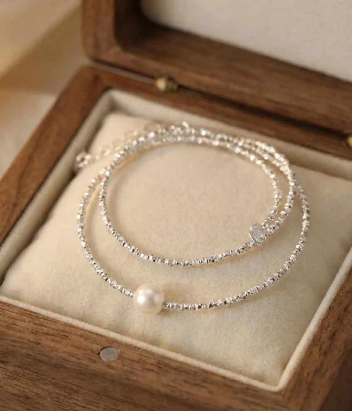 A delicate silver-plated necklace with a shimmering, textured chain and a single, central white pearl, displayed inside a wooden jewelry box on a soft cream-colored cushion.