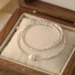 A delicate silver-plated necklace with a shimmering, textured chain and a single, central white pearl, displayed inside a wooden jewelry box on a soft cream-colored cushion.