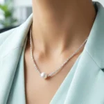 A delicate silver-plated necklace with a shimmering, textured chain and a single, central white pearl, displayed inside a wooden jewelry box on a soft cream-colored cushion.