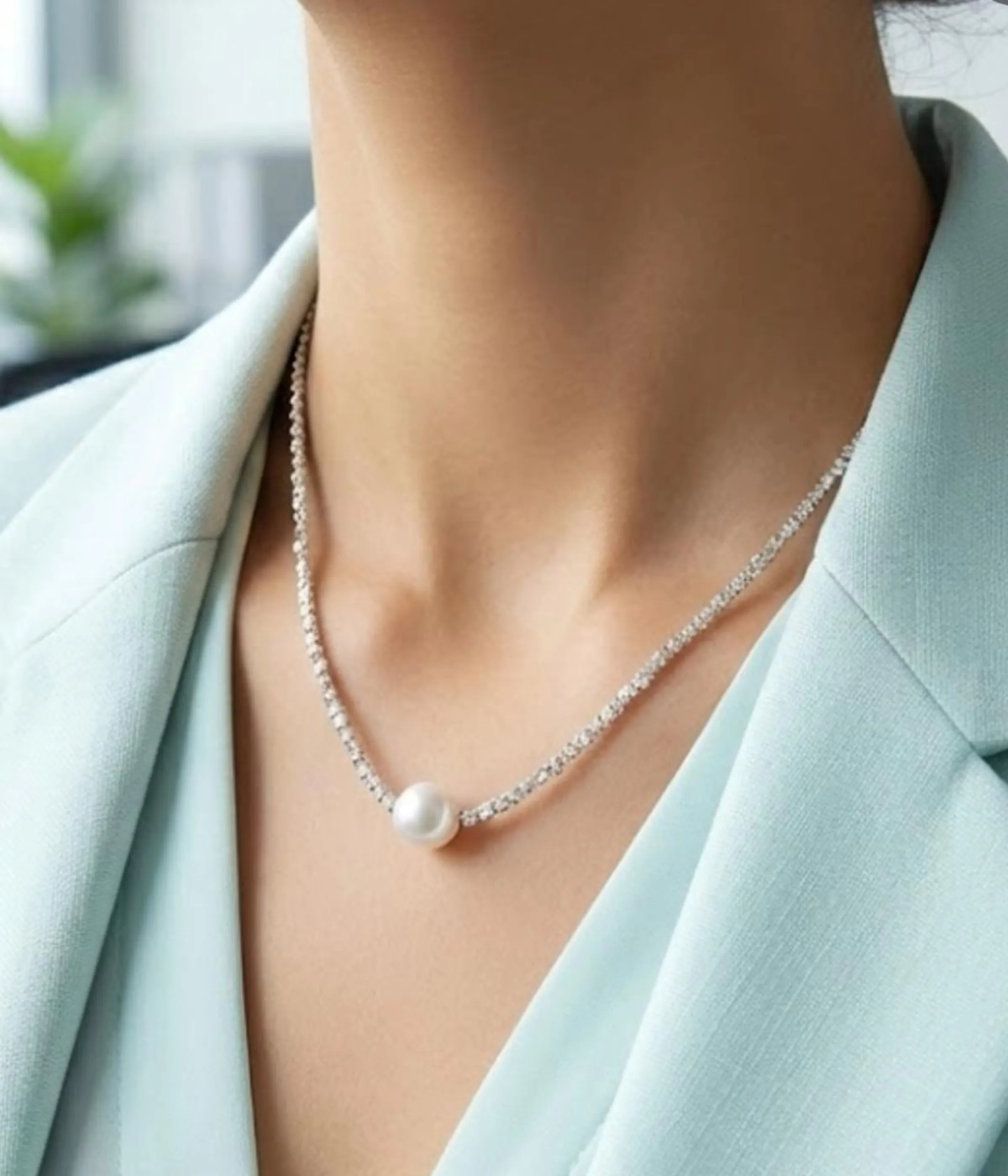 A delicate silver-plated necklace with a shimmering, textured chain and a single, central white pearl, displayed inside a wooden jewelry box on a soft cream-colored cushion.