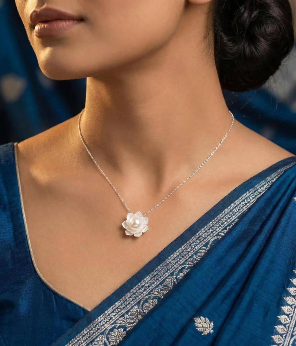 A close-up shot of a silver-plated lotus flower pendant featuring intricate, brushed-metal petals layered around a single, luminous white pearl, displayed on a soft cream jewelry cushion.