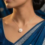 A close-up shot of a silver-plated lotus flower pendant featuring intricate, brushed-metal petals layered around a single, luminous white pearl, displayed on a soft cream jewelry cushion.